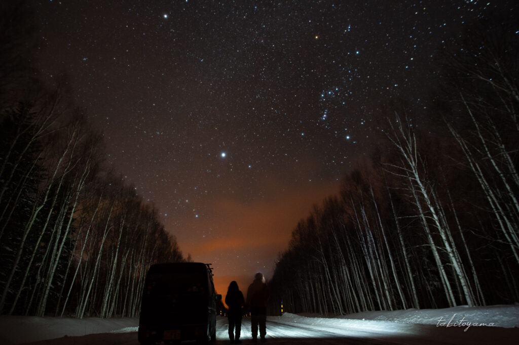 真冬の北海道で車から降りて満点の星空を眺める夫婦