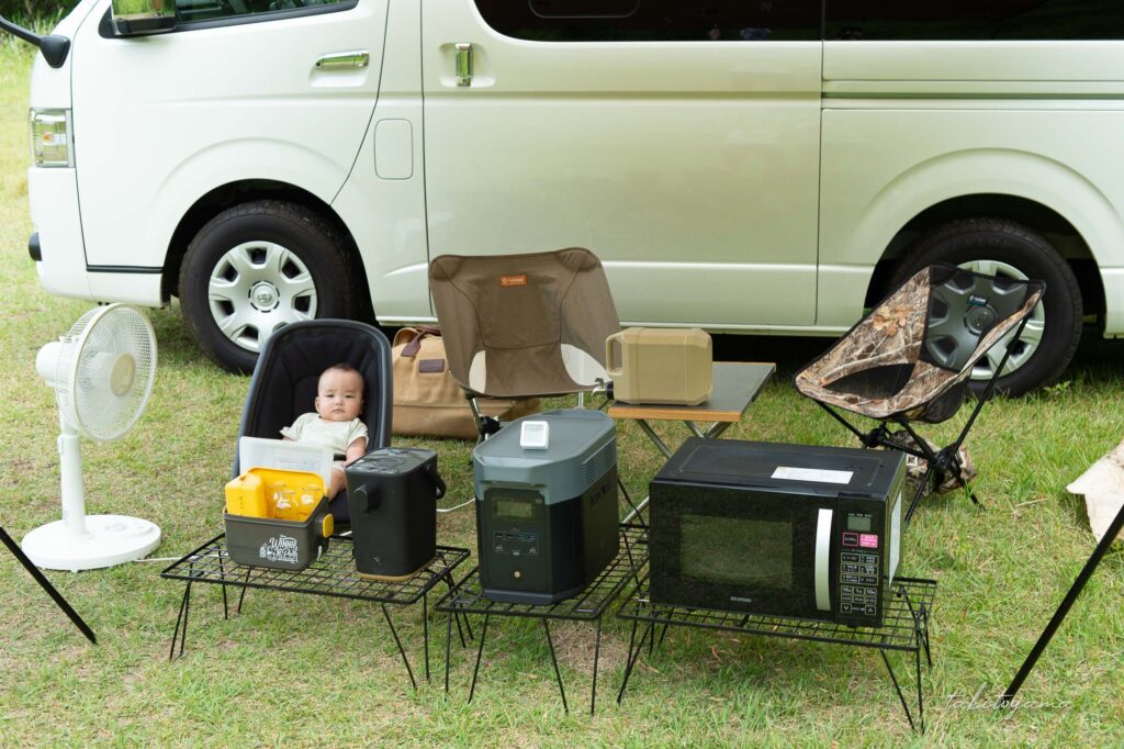 ハイエースを背景に家電がたくさん並べられたそばで扇風機で涼む息子