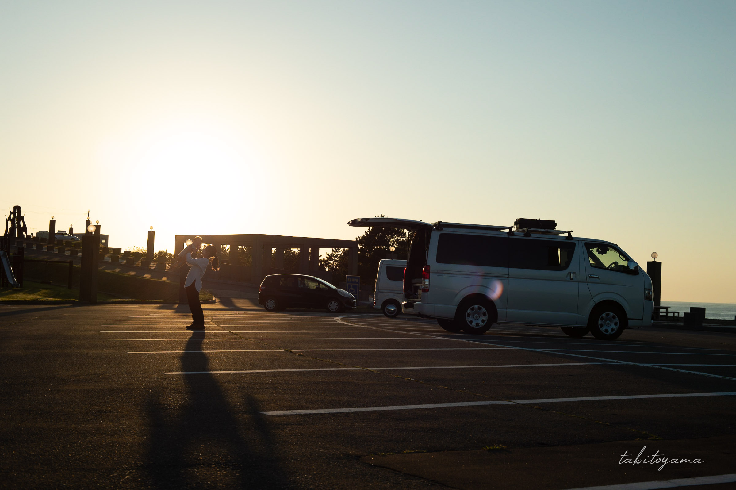 夕陽を背景にバックドアが開いたハイエースと妻に抱き上げられた息子
