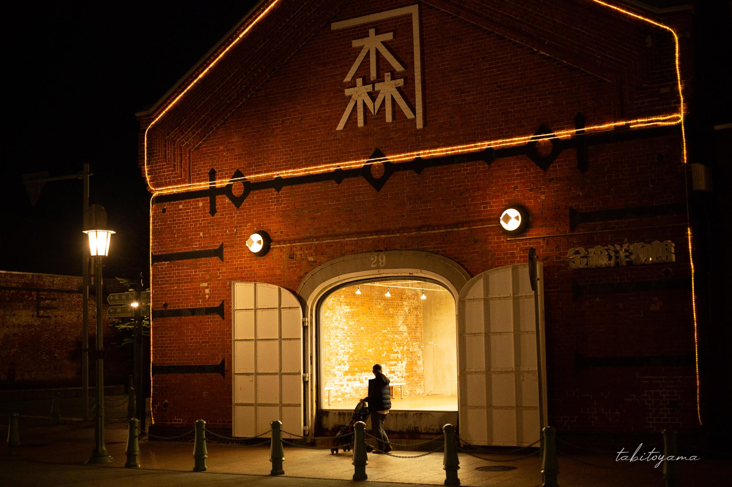 夜の金森赤れんが倉庫前をベビーカーを押しながら歩く妻、窓からの灯りに照らされる