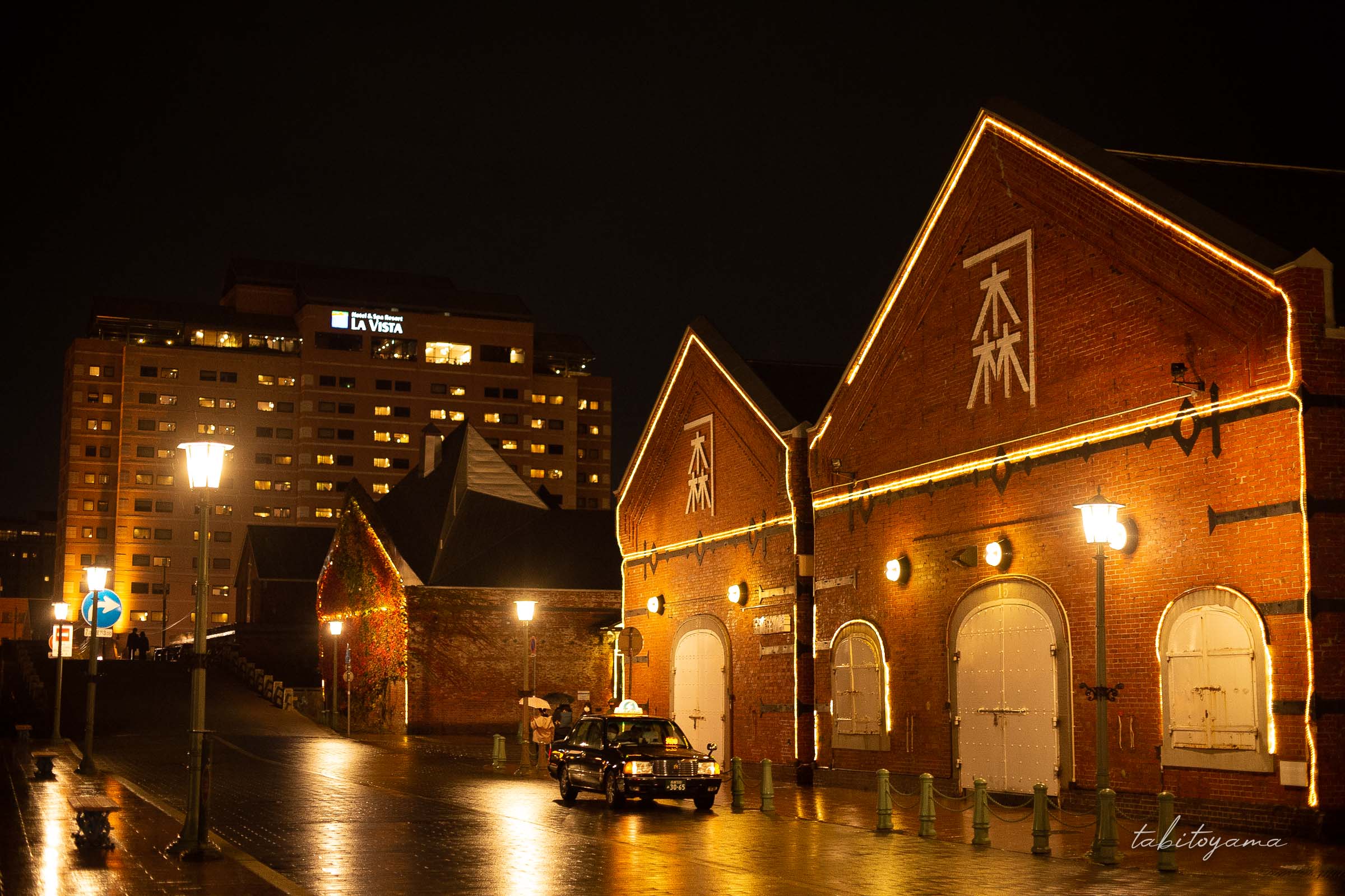 夜金森赤れんが倉庫軍前に停車するタクシー、濡れた路面に街灯が反射し奥にはラビスタ函館ベイが見える