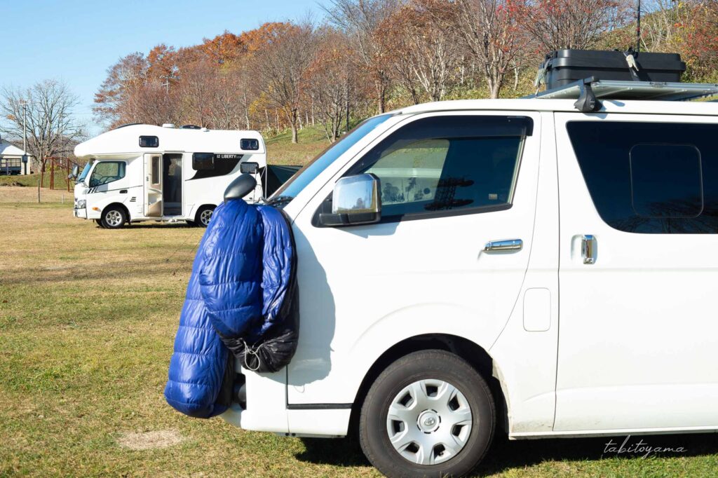 ハイエースのミラーに寝袋が干されている側面からの眺め、奥には大型のキャンピングカー