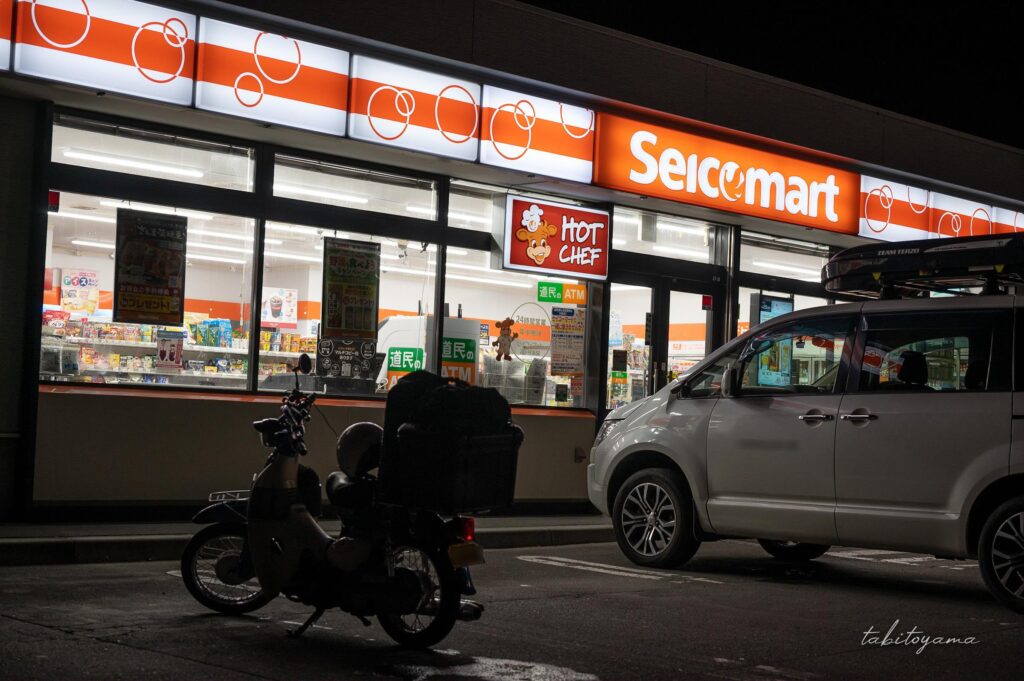 夜のセイコーマートのオレンジ色の看板が光る。駐車場には荷物を積載したバイクが停まる。