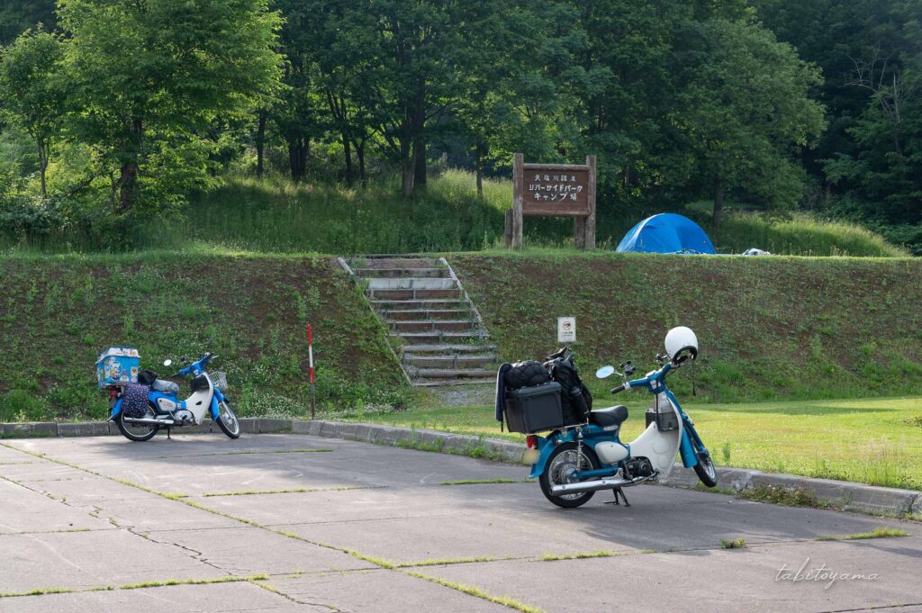 天塩川リバーサイドパークキャンプ場の駐車場。カブが二台駐車されていて、奥の階段付近には看板とテントが設営されている。