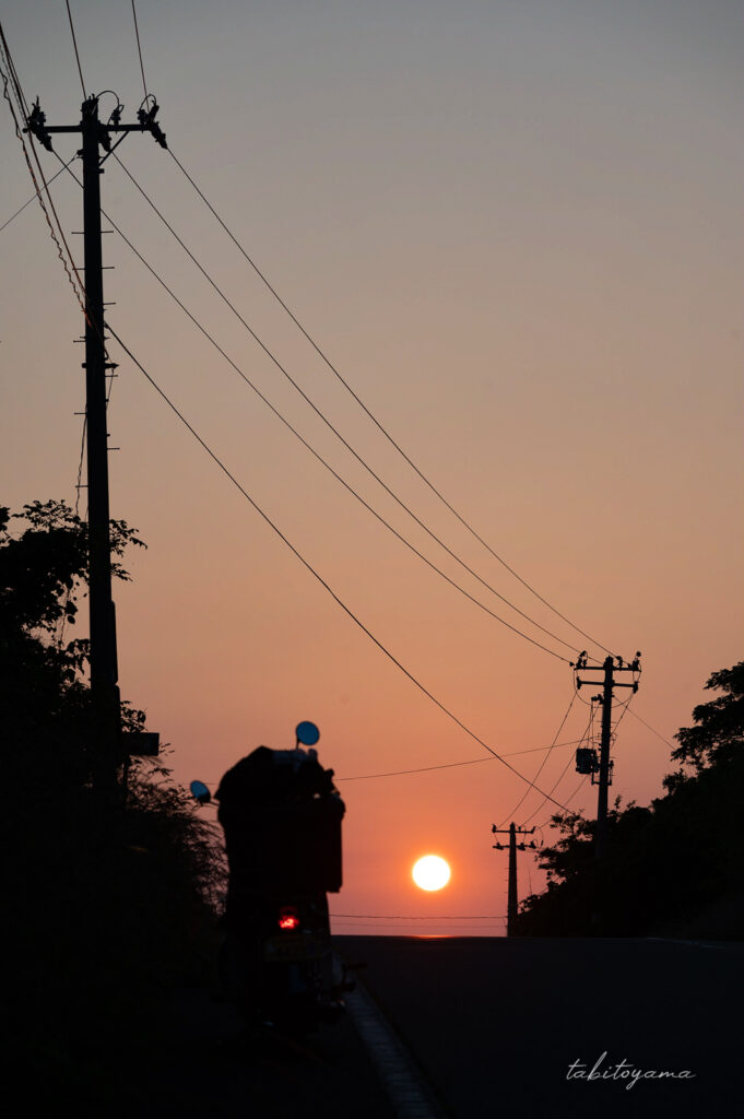坂道を登った先に見える丸い夕日、手前にはシルエットになった電柱とリトルカブ