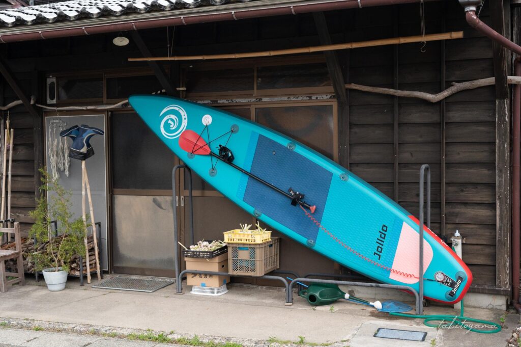 松ヶ崎集落の落ち着いた外観の建物に鮮やかなターコイズブルーのSUPが立て掛けられている