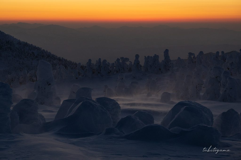 オレンジ色の帯状に染まる夕焼けの空と暗い樹氷原を通り抜ける雪の舞
