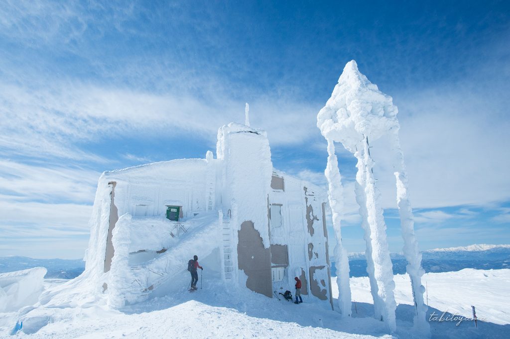 雪に覆われた刈田岳レストハウスの影で休憩する登山者の姿