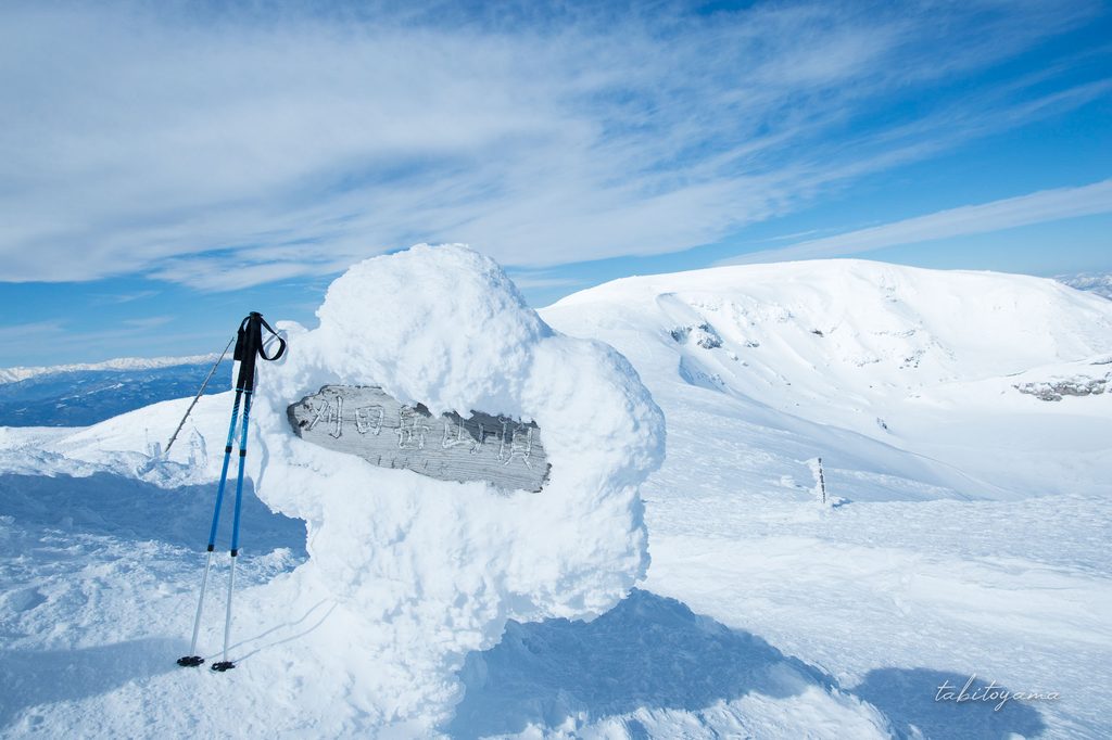 雪に覆われた刈田岳山頂標識に立てかけられたトレッキングポール