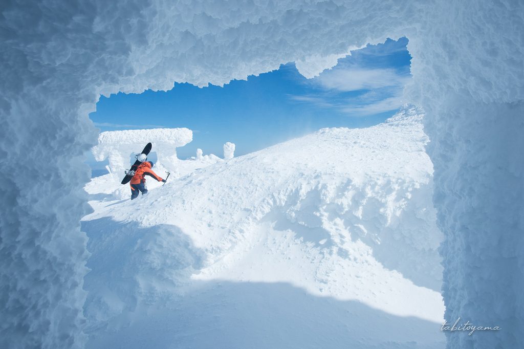 雪に覆われた刈田峰神社の本殿の内部から外を眺めるとボードを担いだ登山者