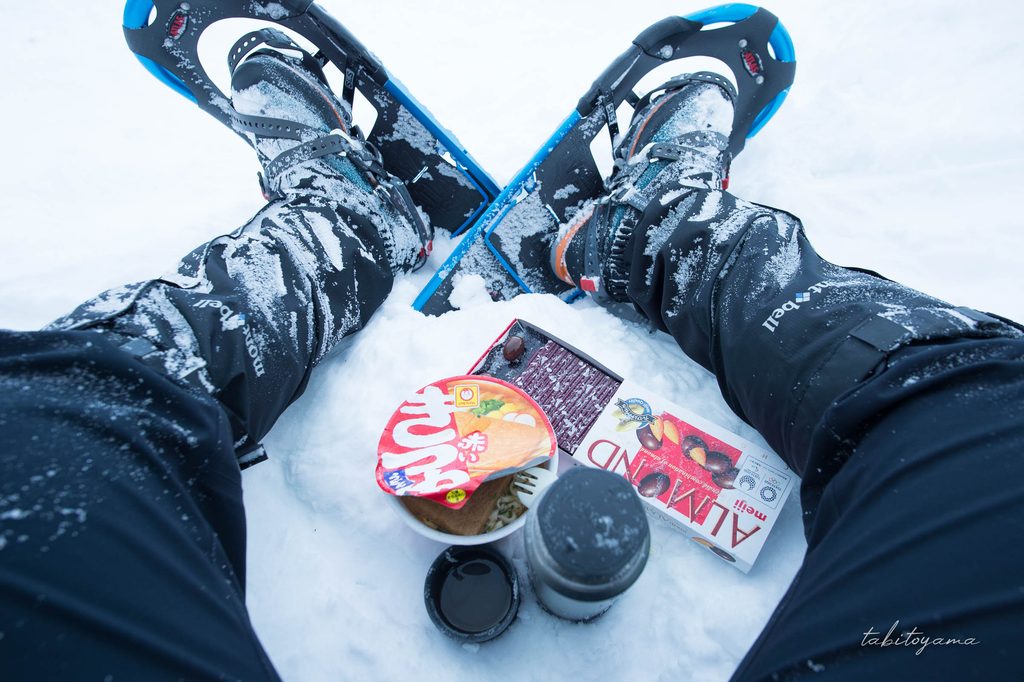 雪の上に腰掛けてスノーシューを履いた両足の中に保温ボトルとカップ麺などが広げられた様子
